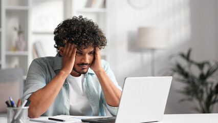 A young Indian male businessman is sitting at a desk trying to work, checking schedules and getting frustrated that something is not working out. Headache, fatigue, burnout.