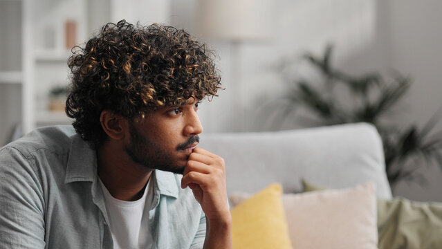 Bored millennial generation man sitting on sofa, feeling lonely at home. Unhappy young male suffering from negative thoughts, having depressive mood or problems in relations, regretting mistakes.