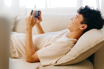 Relaxed young man playing mobile games on smartphone while lying on cozy sofa in bright room with natural light Comfort and leisure concept for home entertainment