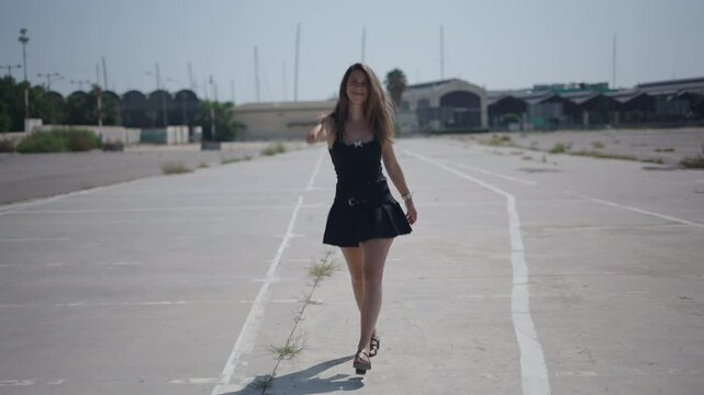 young girl caucasian woman in short black dress walking facing camera in an industrial area on the outskirts near the port making the OK gesture with thumbs up