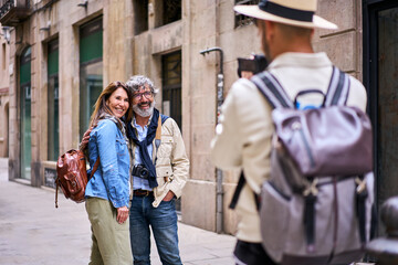 Young male photographer taking holiday photo of happy elderly tourist couple posing smiling on city...