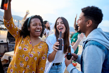 Three diverse young excited friends dancing together using beer bottles like a microphone at rooftop summer party. Multiracial people having fun drinking alcohol celebrating live music terrace