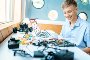 Blonde hair schoolboy in blue shirt watching motherboard while considering and smile in STEM class. On table put laptop, controller, electric wire, battery charger, and robotic vehicle. Edification.