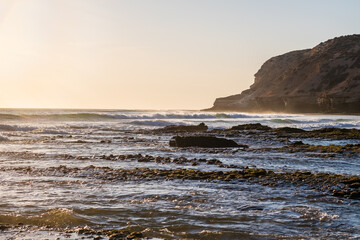 La Source beach in Taghazout near Agadir, Morocco at sunset. Rocky coastline and splashing waves of Atlantic ocean attract surfers and tourists. Beautiful tranquil water background. Famous surf spot