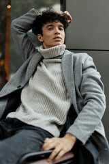 Fashionable young man in gray sweater and black pants sitting on a chair with hands on head, looking at camera