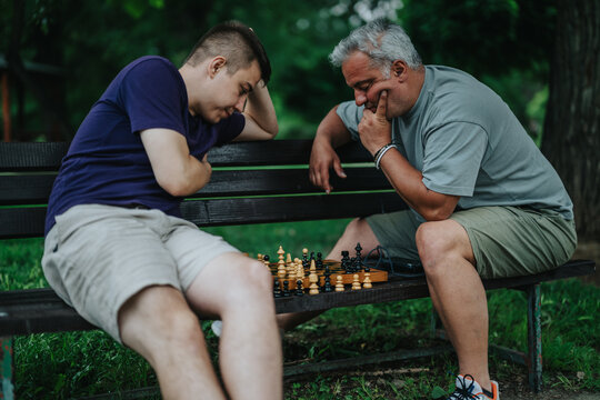A father and son deeply focused on a chess game while sitting on a park bench. The image captures concentration and bonding in an outdoor setting.