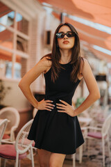 Gorgeous woman posing confidently outdoors wearing a stylish black dress and sunglasses. Urban setting with colorful parasols and outdoor seating. Fashion and confidence concept.