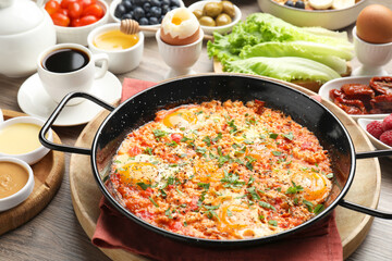 Tasty shakshouka in frying pan and other meals served for breakfast on wooden table