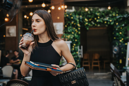 Fashionable young woman sipping iced coffee outside a stylish cafe while carrying notebooks and a black handbag.