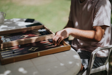 Person playing an engaging game of backgammon outdoors, enjoying a peaceful afternoon under the shade. Focused on strategy and leisure in a natural setting during a sunny day.