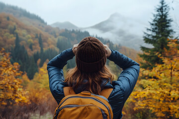 Woman hiker adjusting her hat while standing in front of misty autumn landscape with vibrant fall trees and fog-covered mountains