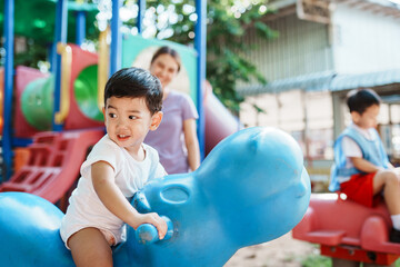 A young Asian boy, happy playing as rides a rocking horse at the playground, his mother standing close by to watch over him and ensure his safety during playtime.