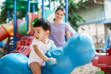 Asian boy, playing as rides a rocking horse at the playground, his mother standing close by to watch over him and ensure his safety during playtime.