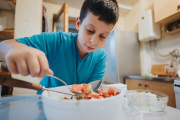 A young boy in a blue shirt is focused on preparing a fresh vegetable salad in a home kitchen, promoting healthy eating and culinary skills in children.