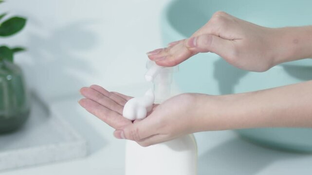 Close-up of foam taken from unlabeled product bottle on white table with bathroom background. A woman's hands are gathering foam to prepare for washing her face or taking a bath