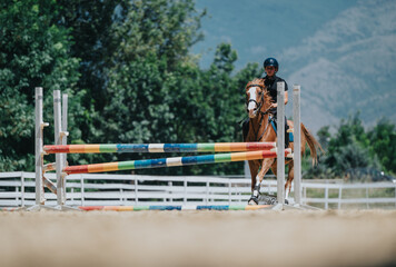 Equestrian rider in helmet jumping horse over colorful barrier in outdoor arena with scenic mountain background.