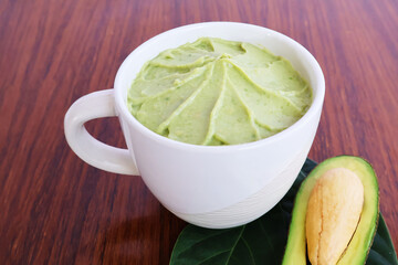 Pureed avocado pulp in a cream-colored ceramic bowl on a wooden table with leaves and halved ripe fruit.

