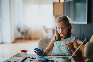 Fototapeta premium A focused young girl paints with watercolors at home, expressing creativity and concentration during an art activity.
