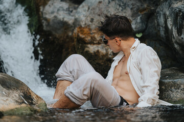 A young man in casual wear relaxes by a waterfall, sitting on rocks with rolled-up pants and a casual shirt.