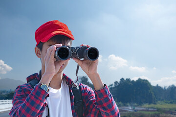 Obraz premium Asian boy wearing a red cap and plaid shirt, looking through binoculars while standing on a bridge over a river. He’s enjoying the natural surroundings and observing wildlife with curiosity.