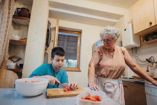 A grandmother supervises her grandson as he chops vegetables in their cozy kitchen. The scene captures generational bonding and shared culinary experiences.