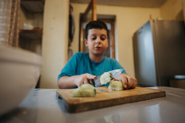 A young boy in a blue shirt carefully cuts vegetables in a cozy kitchen. Learning basic cooking skills, he uses a knife with focus and care.