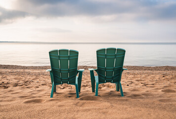 Two teal adirondack chairs on sandy beach with view of lake in summer.