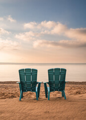 Two teal adirondack chairs on sandy beach with view of lake in summer.
