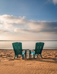 Two teal adirondack chairs on sandy beach with view of lake in summer.