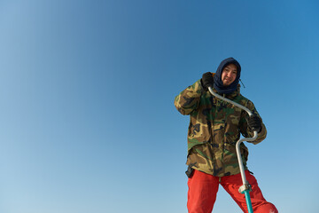 A warmly dressed Asian guy drills a hole in the ice on winter fi