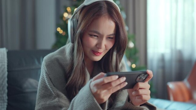 Close up of serious young asian woman wearing headphone playing game online with mobile phone at home living room decorated for Christmas, MOBA MMORPG	