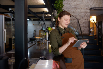 Young female barista entering order on mobile tablet standing at counter desk