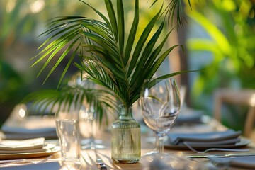 Outdoor dining scene with rich garden backdrop. White tablecloth features palm fronds centerpiece, silverware. Perfect for festive banquet or special occasion. Elegant atmosphere with natural beauty.
