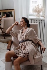 A woman is sitting in a chair wearing a tan jacket and a leopard print dress. She is wearing a belt...