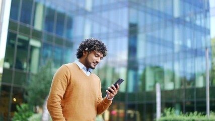 Handsome happy businessman in casual walking near modern business office building hold smartphone in hands. Young adult Man entrepreneur employee using read a mobile phone outside urban lifestyle city