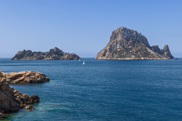 Fototapeta premium Clear view of Es Vedra and Es Vedranell islands off the coast of Ibiza, Spain, with a sailboat in the foreground, highlighting the rugged rocky formations and the deep blue Mediterranean waters