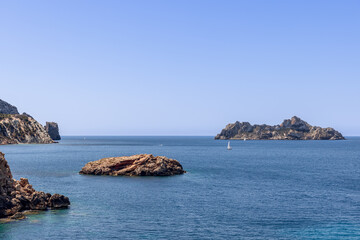 Fototapeta premium Es Vedranell and other rocky islets near Ibiza, Spain, lie in calm blue waters. A sailboat sails freely, embodying the sense of freedom in this tranquil seascape