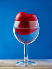Red heart in a wine glass with water on wooden surface, blue color background. Optical illusion, Concept of love and romance. Copy space.
