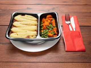 Cottage pie meal with peas and carrots in dark plastic container from microwave on white plate and wooden table. High quality meal for quick dinner at office or home. Popular British style food.