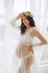 Smiling pregnant woman in white polka dot peignoir and gypsophila wreath posing near window in the studio. Beautiful pregnancy