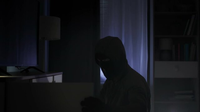robbery, a thief in black clothes and a balaclava rummages through other people's things in search of valuables at night in an apartment