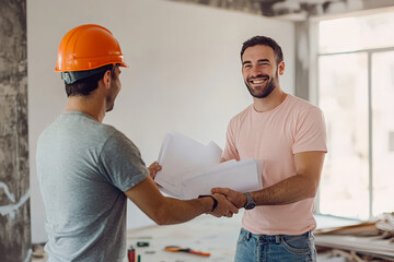 A smiling homeowner shakes hands with a construction worker while reviewing blueprints during a home renovation project.