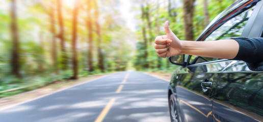 Naklejka premium A hand with a thumbs up gesture is sticking out of a car window on a road with lush green trees on either side, sunshine in the background.