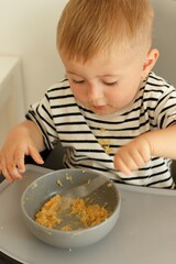 Little serious child eats independently with a spoon while sitting on a high chair.