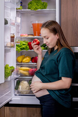 Pregnant woman is eating an apple near the refrigerator