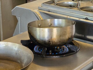 A large stainless steel pot heating over a gas stove in a commercial kitchen. The flames under the pot indicate active cooking or preparation for boiling.

