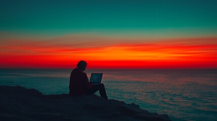 Silhouette of a person working on a laptop with a beautiful orange sunset over the ocean.