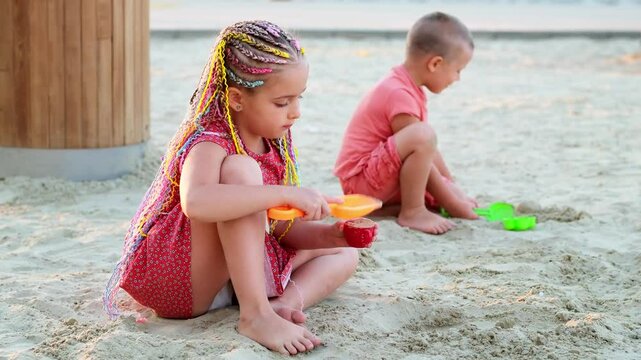 a child plays in the sandbox on the playground in summer, a little girl with bright pigtailed hair plays with sand on the beach and smiles, children's holidays