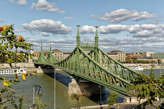 A majestic green iron bridge spans the wide river under a bright cloudy sky, framed by foreground leaves. These iconic architectural achievements inspire global travel and connect vibrant communities.