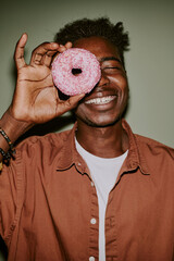 Young man holding pastel pink doughnut to hide his eye while posing for photo